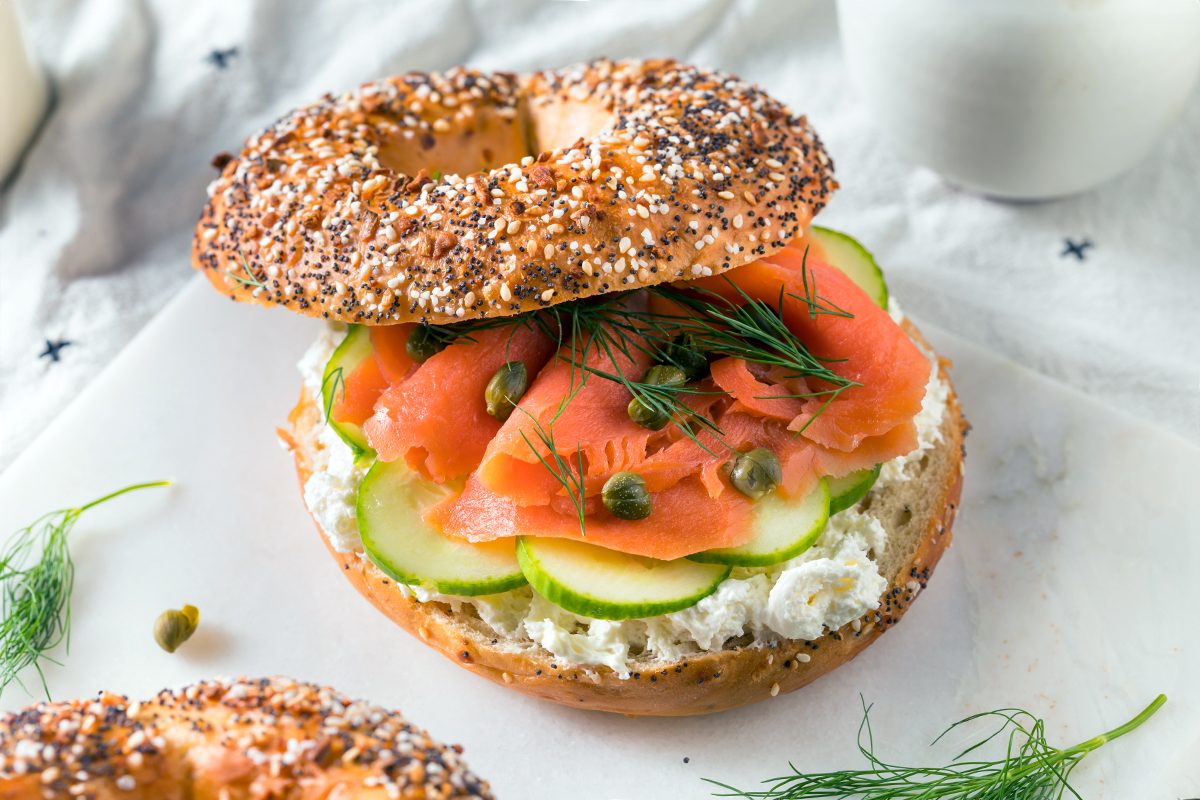 An everything bagel sandwich with cream cheese, smoked salmon, cucumber slices, capers, and fresh dill on a white surface with herb garnish nearby.